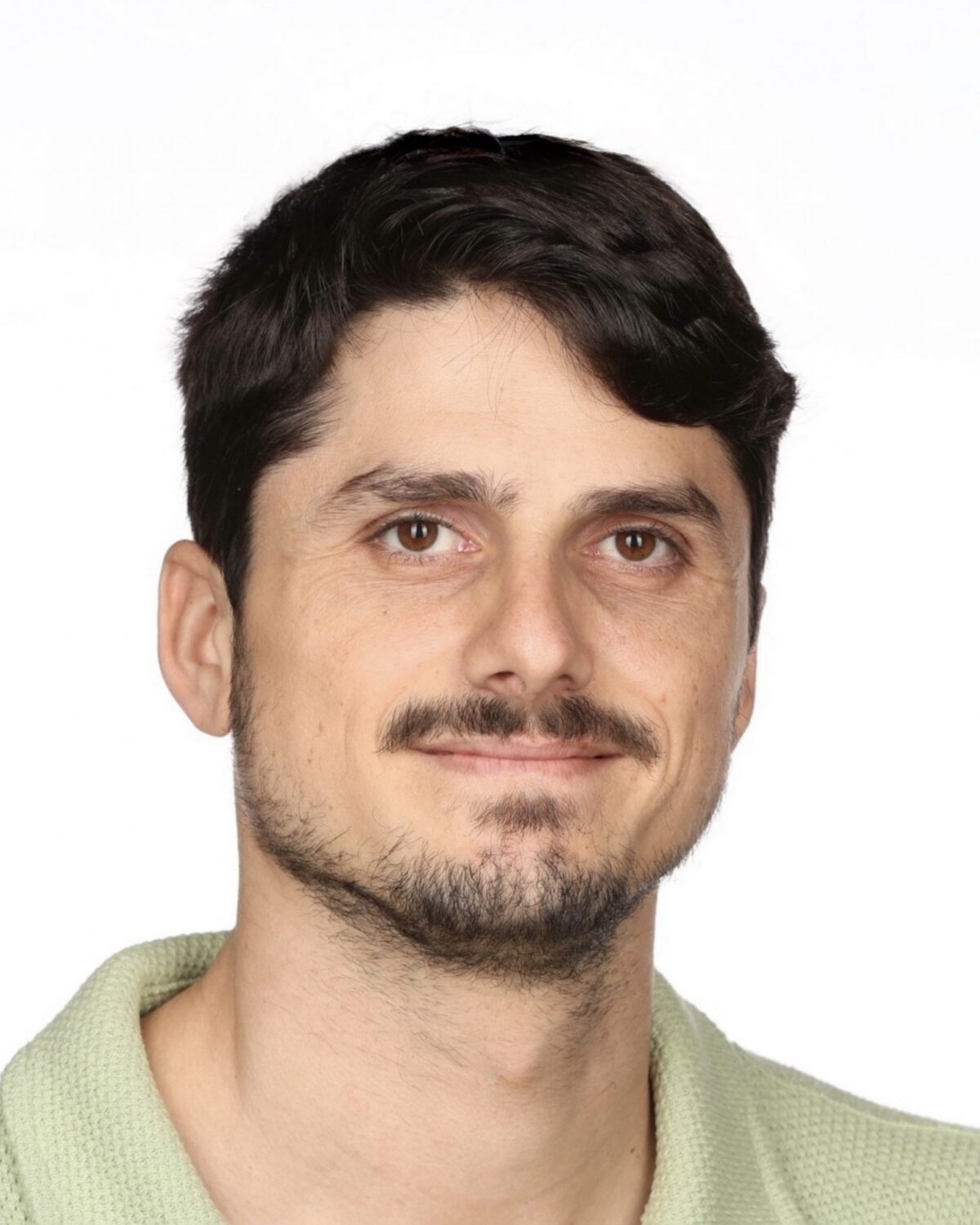 A man with short dark hair, a trimmed beard, and mustache smiles slightly. He is wearing a light green collared shirt against a plain white background.