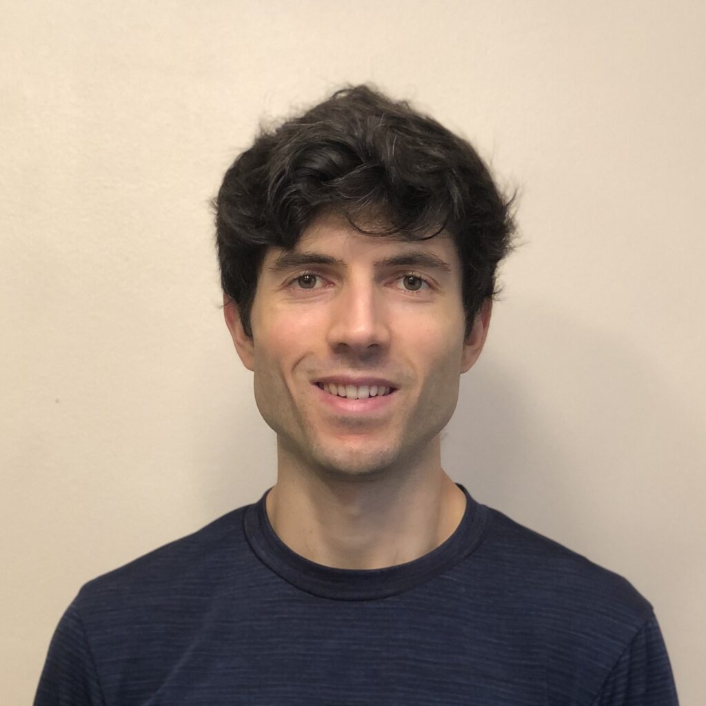 A young man with short, wavy dark hair smiles at the camera. He wears a dark navy shirt and stands in front of a plain, light-colored background.