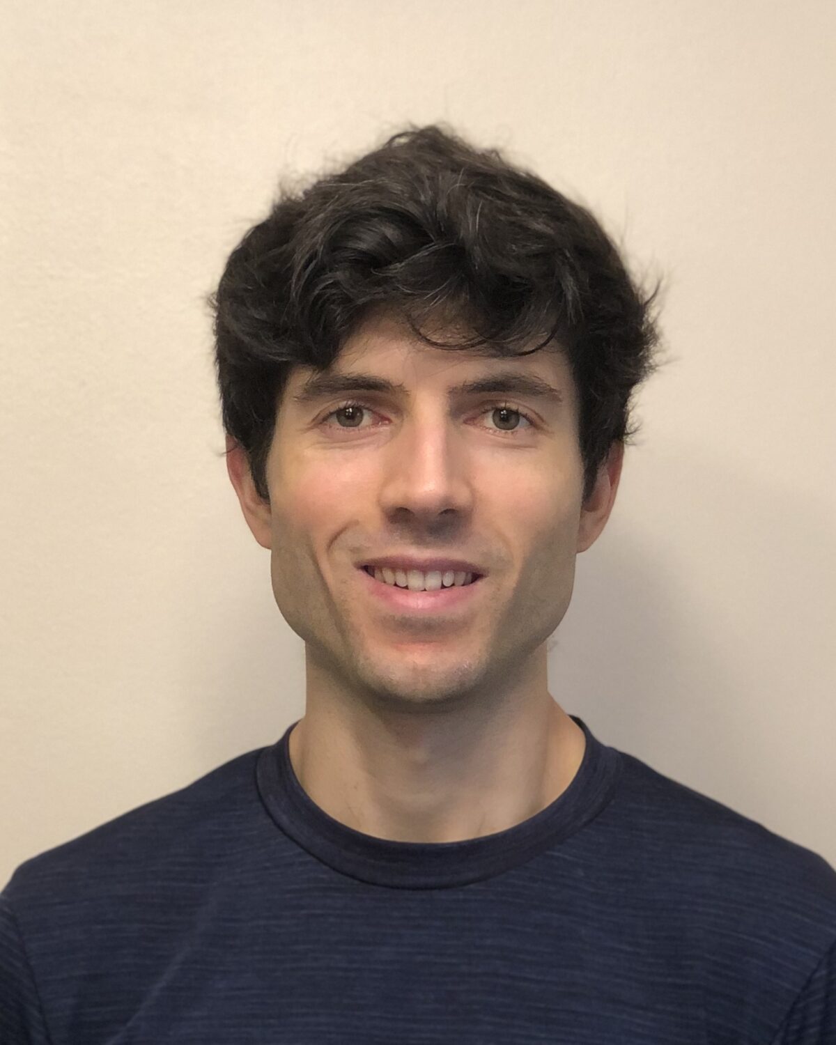 A young man with short, wavy dark hair smiles at the camera. He wears a dark navy shirt and stands in front of a plain, light-colored background.