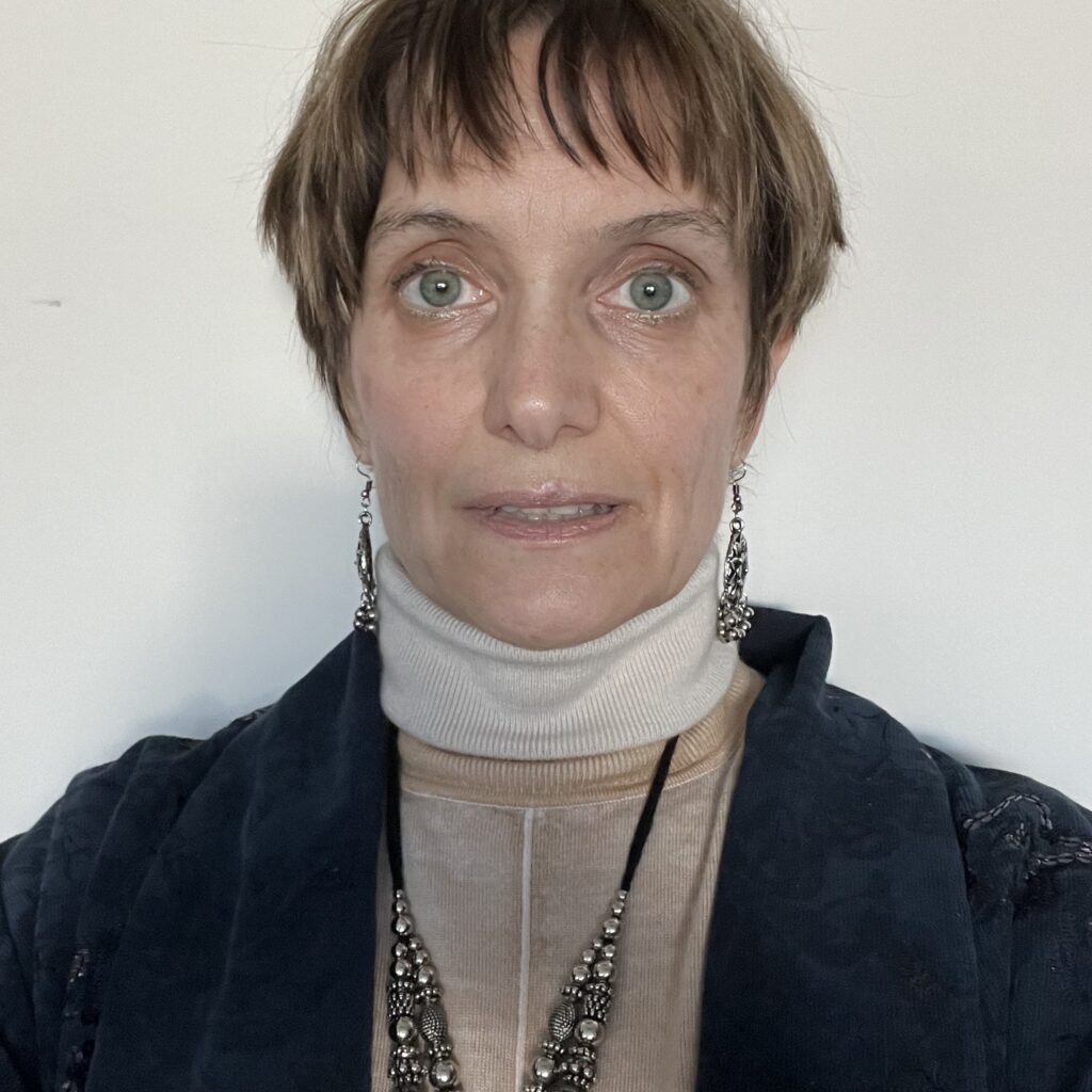 A person with short brown hair and green eyes wears a tan turtleneck, a dark jacket, and beaded jewelry, posing in front of a plain white background.