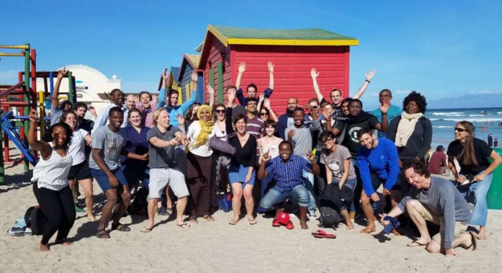 A large, diverse group of people poses and smiles on a sandy beach near colorful huts under a clear blue sky, some raising their hands in excitement. The ocean and distant people are visible in the background.