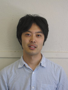 A man with short black hair wearing a light blue, striped button-up shirt stands in front of a plain, off-white wall, looking at the camera with a neutral expression.