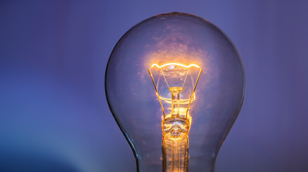 A close-up of a glowing incandescent light bulb against a softly blurred blue and purple background. The filament is clearly visible, radiating warm light inside the transparent glass bulb.