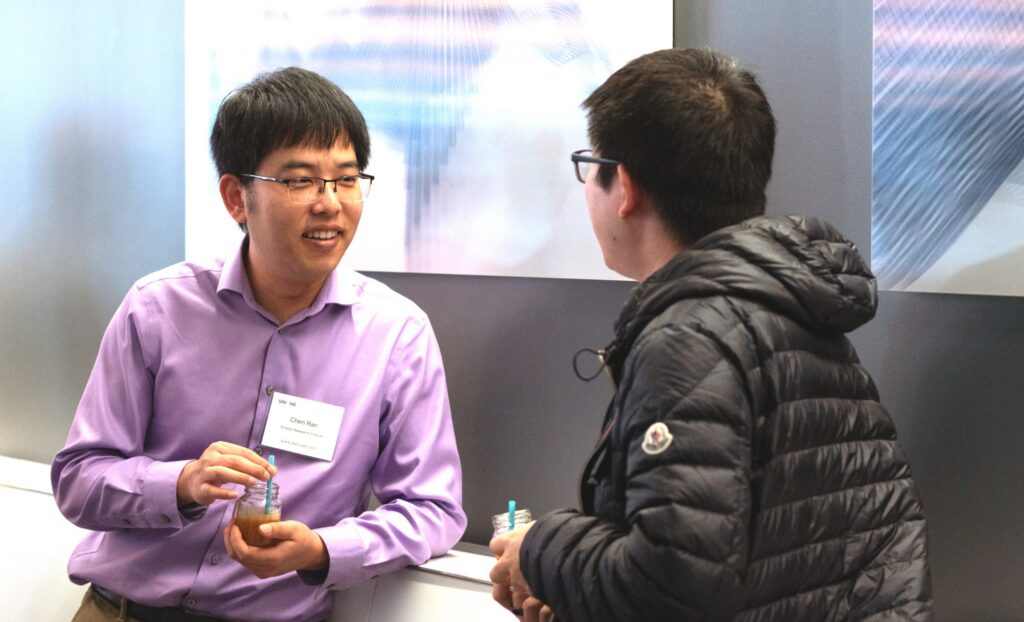 Two people are having a conversation indoors. One wears a purple shirt and name tag, holding a drink. The other, seen from the back, wears glasses and a black jacket. Abstract art is visible on the wall behind them.