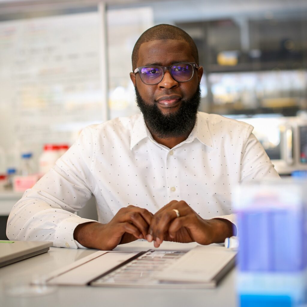A man with glasses and a beard sits at a desk in a lab, wearing a white shirt and looking at the camera. Lab equipment and a whiteboard are visible in the background.