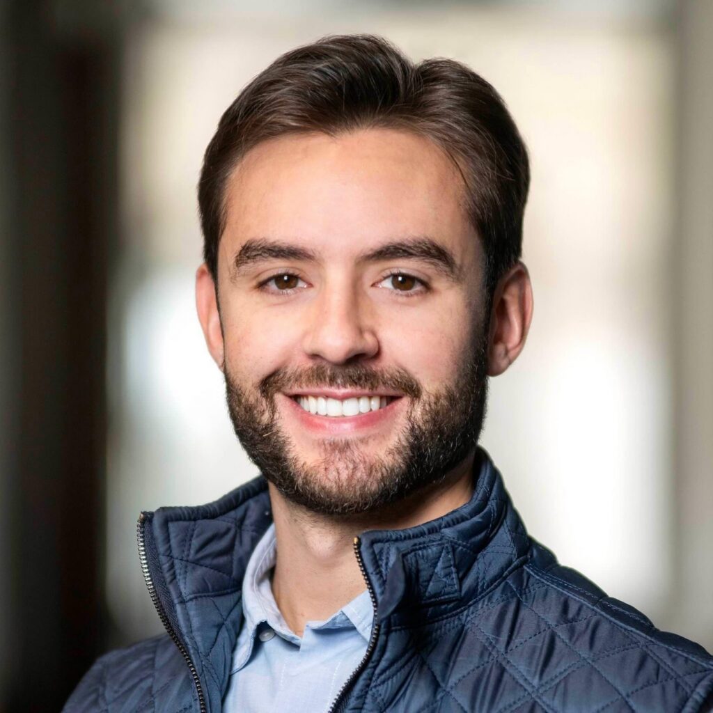 A young man with short brown hair and a beard smiles at the camera. He is wearing a light blue shirt and a dark quilted vest, with a blurred indoor background.
