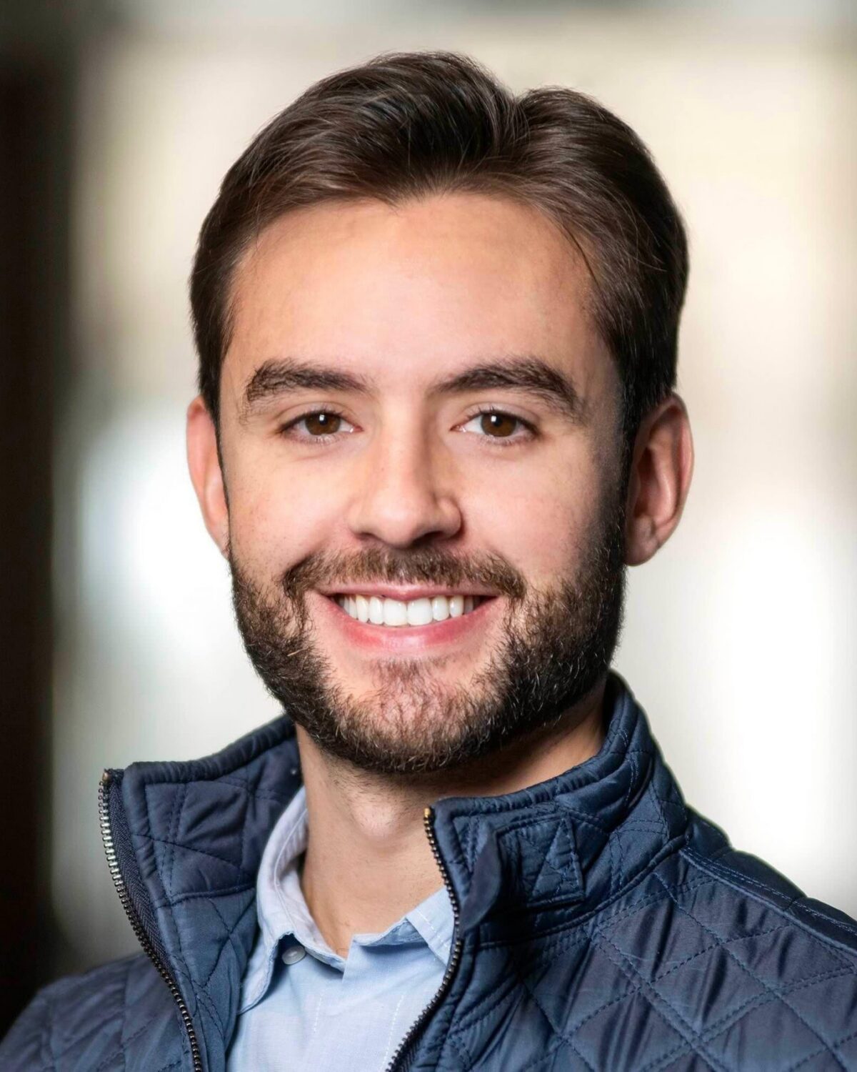A young man with short brown hair and a beard smiles at the camera. He is wearing a light blue shirt and a dark quilted vest, with a blurred indoor background.