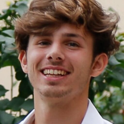 A young person with wavy brown hair smiles at the camera, wearing a white shirt. Green leaves and a beige wall are visible in the background.