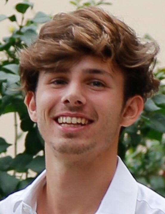 A young person with wavy brown hair smiles at the camera, wearing a white shirt. Green leaves and a beige wall are visible in the background.