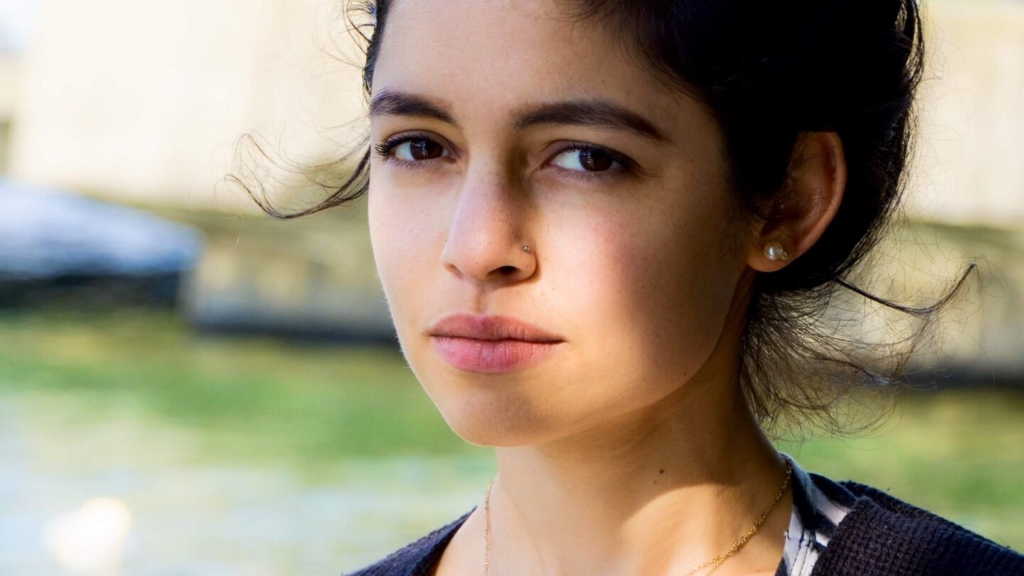 A person with dark hair in a bun, wearing a dark sweater and layered necklaces, stands outdoors near water, looking seriously at the camera.