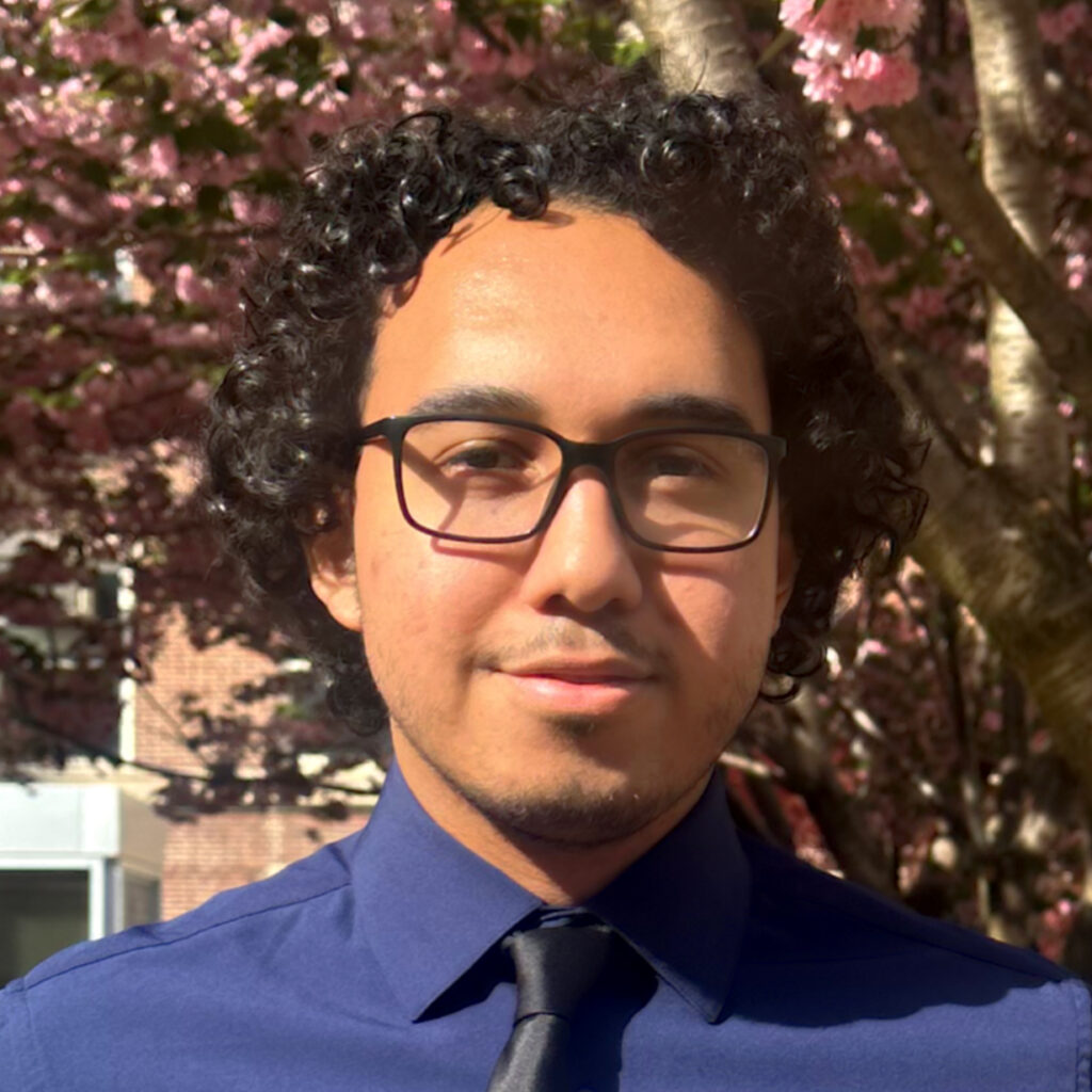 A person with curly dark hair and glasses wearing a blue shirt and black tie stands outside in sunlight, with pink flowering trees in the background.