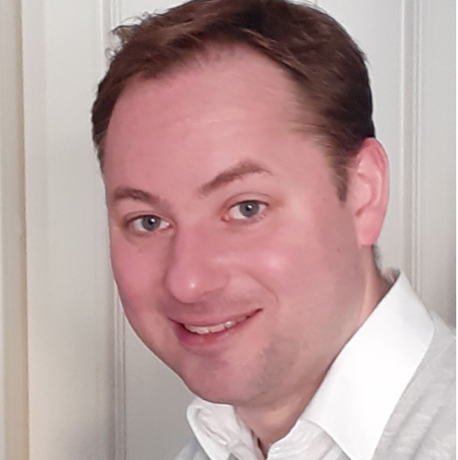 A man with short brown hair and blue eyes is smiling at the camera. He is wearing a white collared shirt and a light gray sweater, standing in front of a white paneled wall.