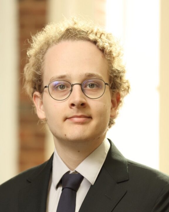 A person with curly blonde hair, glasses, and a suit and tie stands indoors in front of a blurred background with soft light.