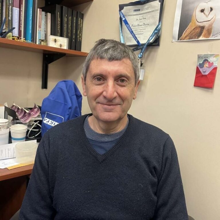 A man with short gray hair and a dark sweater sits in an office, smiling. Behind him are shelves with books, a certificate, a photo of an owl, and a colorful drawing on the wall.