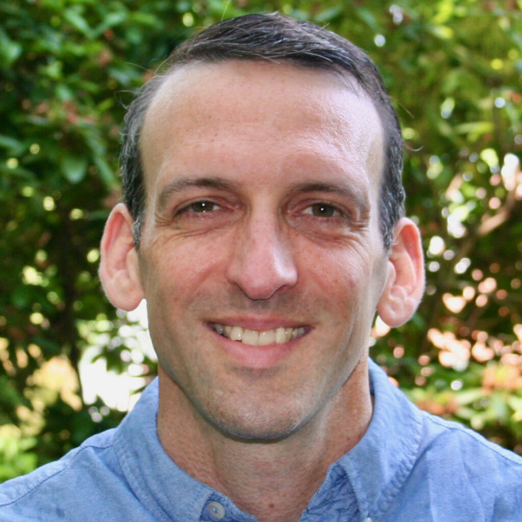 A man with short dark hair and a blue collared shirt smiles at the camera, standing in front of green foliage outdoors.