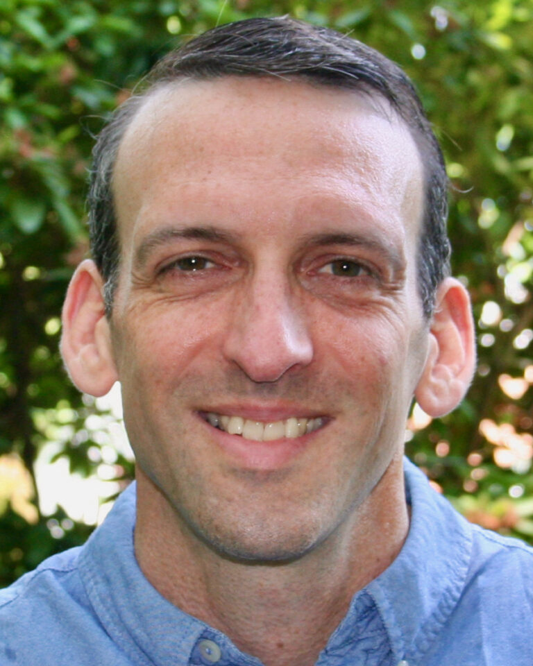 A man with short dark hair and a blue collared shirt smiles at the camera, standing in front of green foliage outdoors.