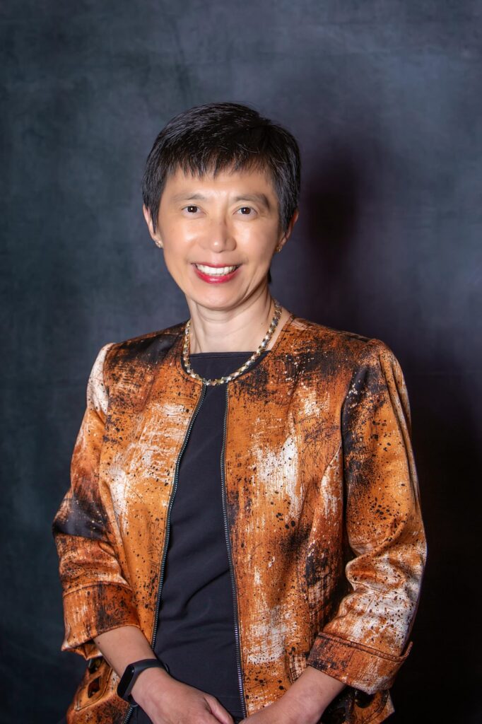A woman with short dark hair smiles at the camera. She wears a black top and a brown, patterned jacket, along with a necklace and a smartwatch. The background is dark and plain.