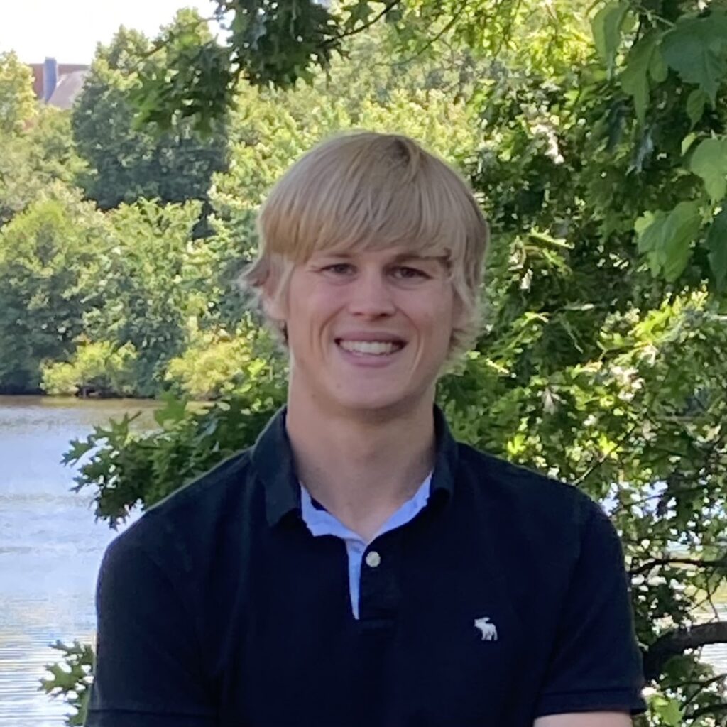 A young man with blond hair, wearing a navy polo shirt, smiles while standing outdoors in front of green trees and a body of water.