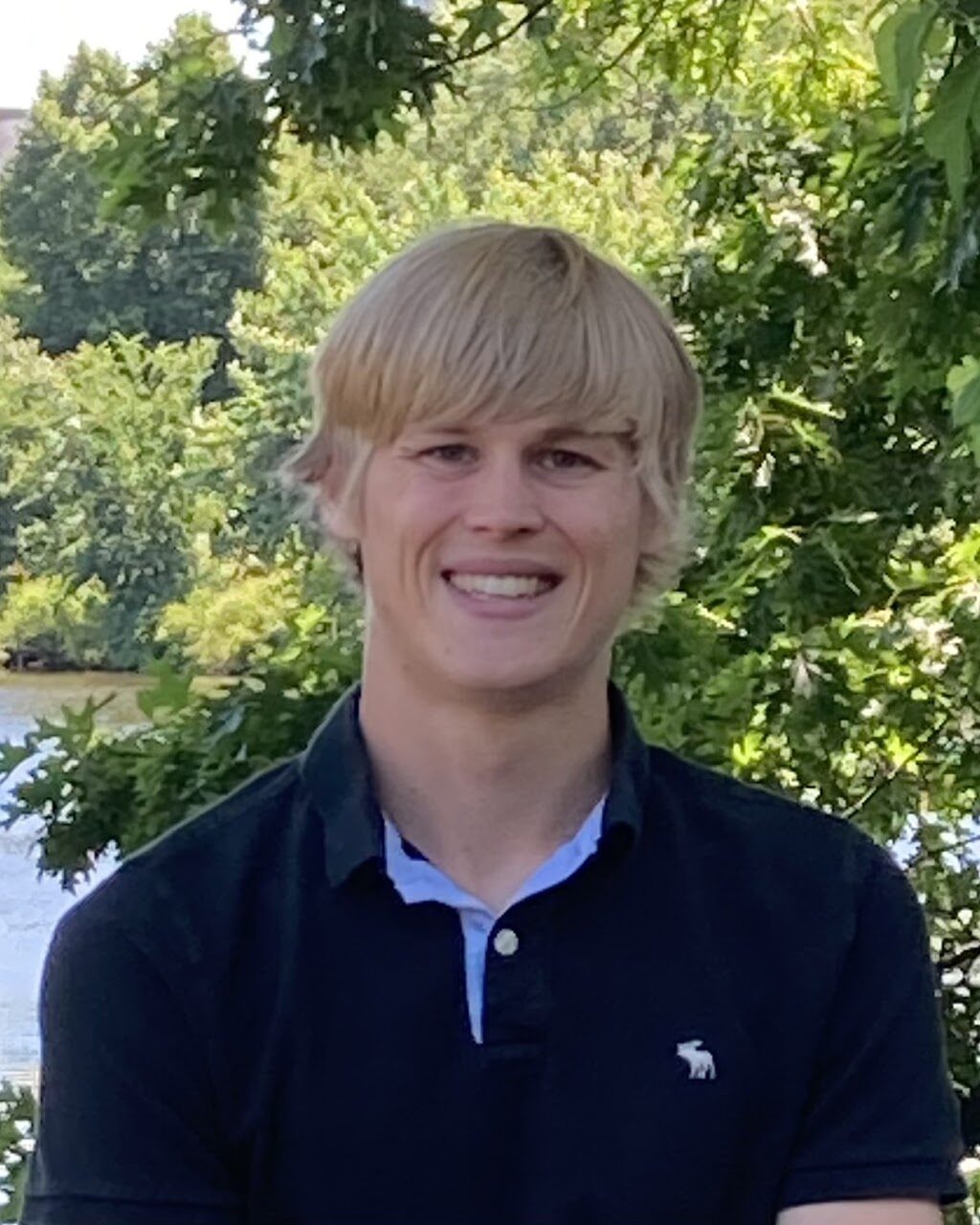 A young man with blond hair, wearing a navy polo shirt, smiles while standing outdoors in front of green trees and a body of water.