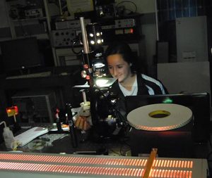 A person in a dark lab sits at a desk, using scientific equipment and computers. The setup includes lights, wires, and illuminated components, indicating a research or experiment environment.