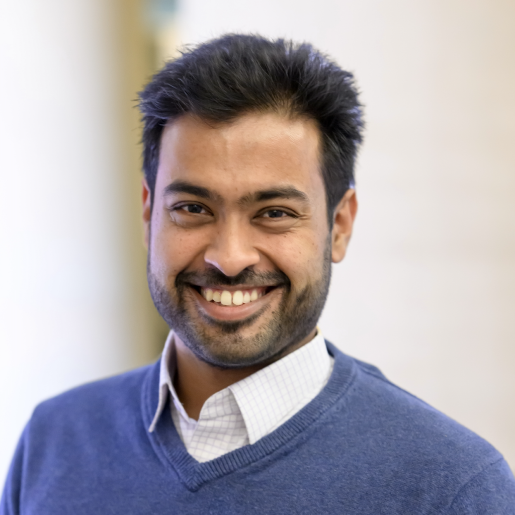 A man with dark hair and a beard smiles at the camera. He is wearing a blue sweater over a collared shirt. The background is softly blurred in neutral tones.