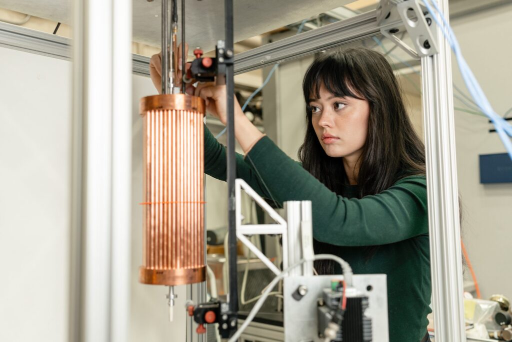 A person with long dark hair adjusts a piece of copper scientific equipment in a lab setting, surrounded by metal structures and wires.