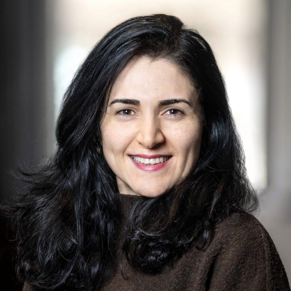 A woman with long dark hair and a brown top smiles at the camera against a softly blurred indoor background.