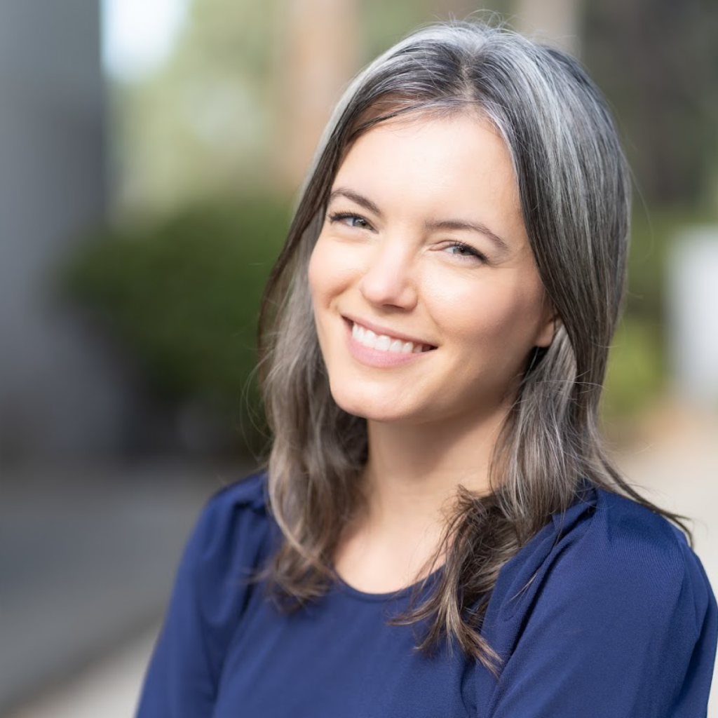 A woman with long brown and gray hair, wearing a navy blue top, smiles outdoors with a blurred green and beige background.
