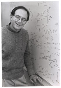 A person wearing glasses and a sweater stands by a whiteboard covered with mathematical equations and diagrams, smiling at the camera.
