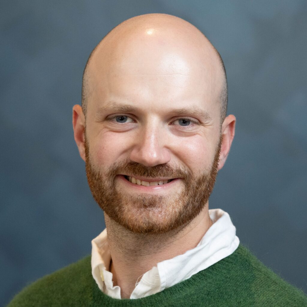 Smiling bald man with a reddish beard wearing a green sweater over a white collared shirt, posed in front of a softly blurred blue-gray background.