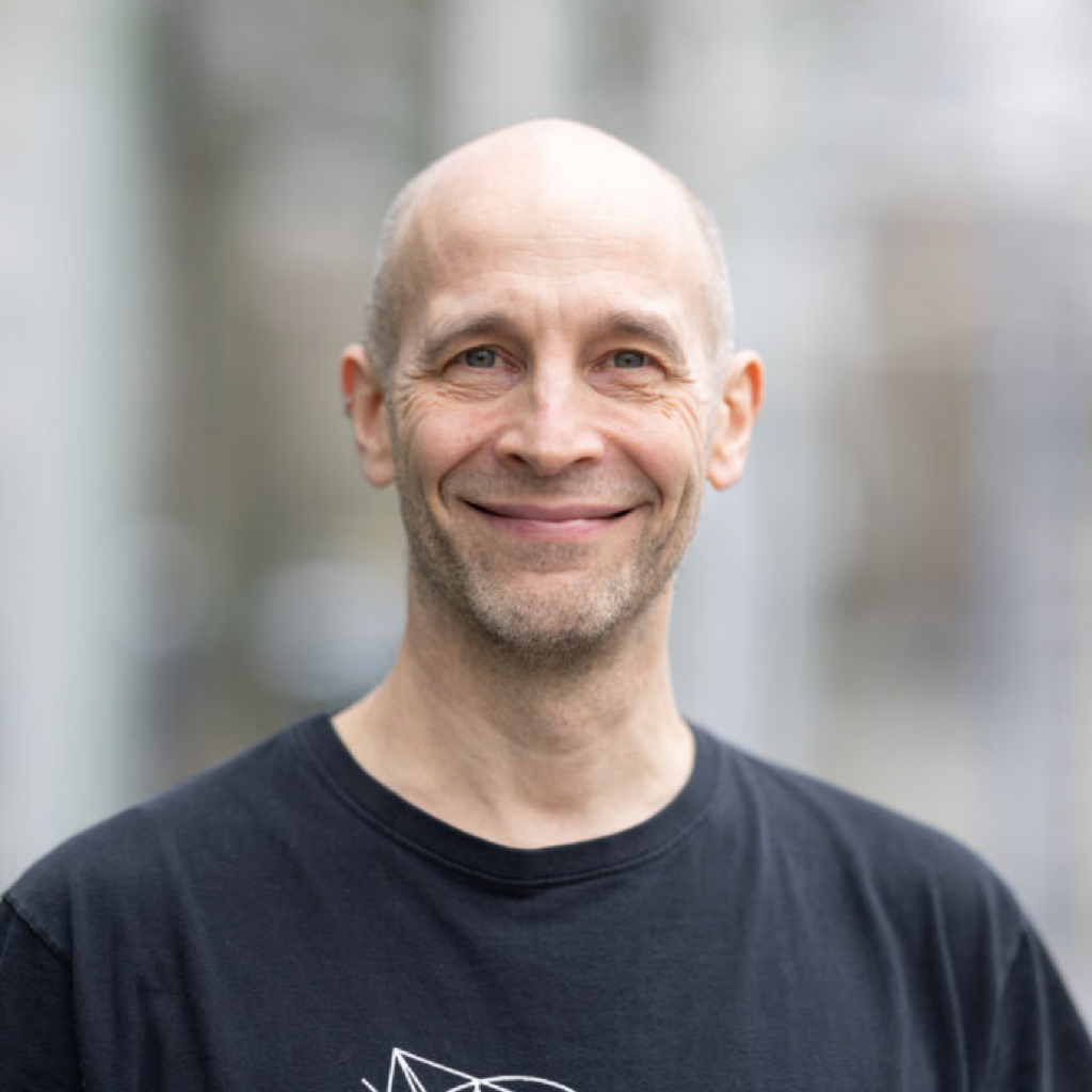 A smiling bald man wearing a black T-shirt stands outdoors with a blurred background.