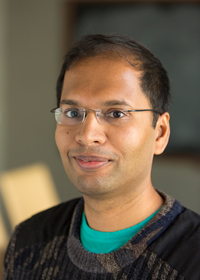A man with short dark hair and glasses, wearing a dark sweater over a green shirt, smiles gently while looking at the camera. The background is softly blurred.