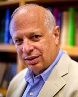 A middle-aged man with a bald head wearing a light-colored blazer and blue shirt, standing in front of a bookshelf filled with books.