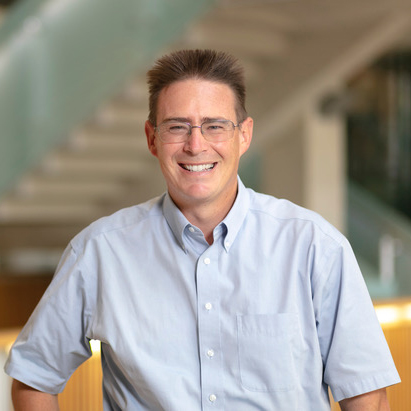 A person wearing glasses and a light blue short-sleeve shirt smiles while standing indoors, with a blurred staircase and modern interior in the background.