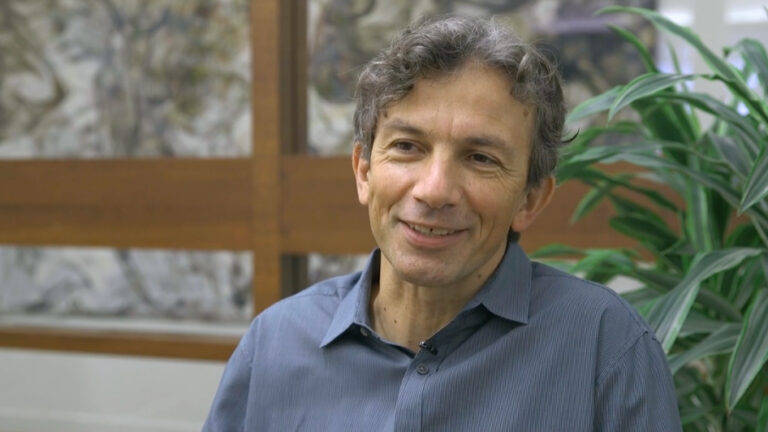 A man with wavy gray hair wearing a gray button-up shirt sits indoors, smiling, with a green plant and wooden paneling in the background.
