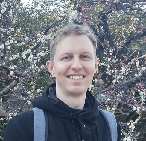 A person with short blond hair, wearing a black jacket and gray backpack, smiles in front of blooming trees with white and pink flowers.