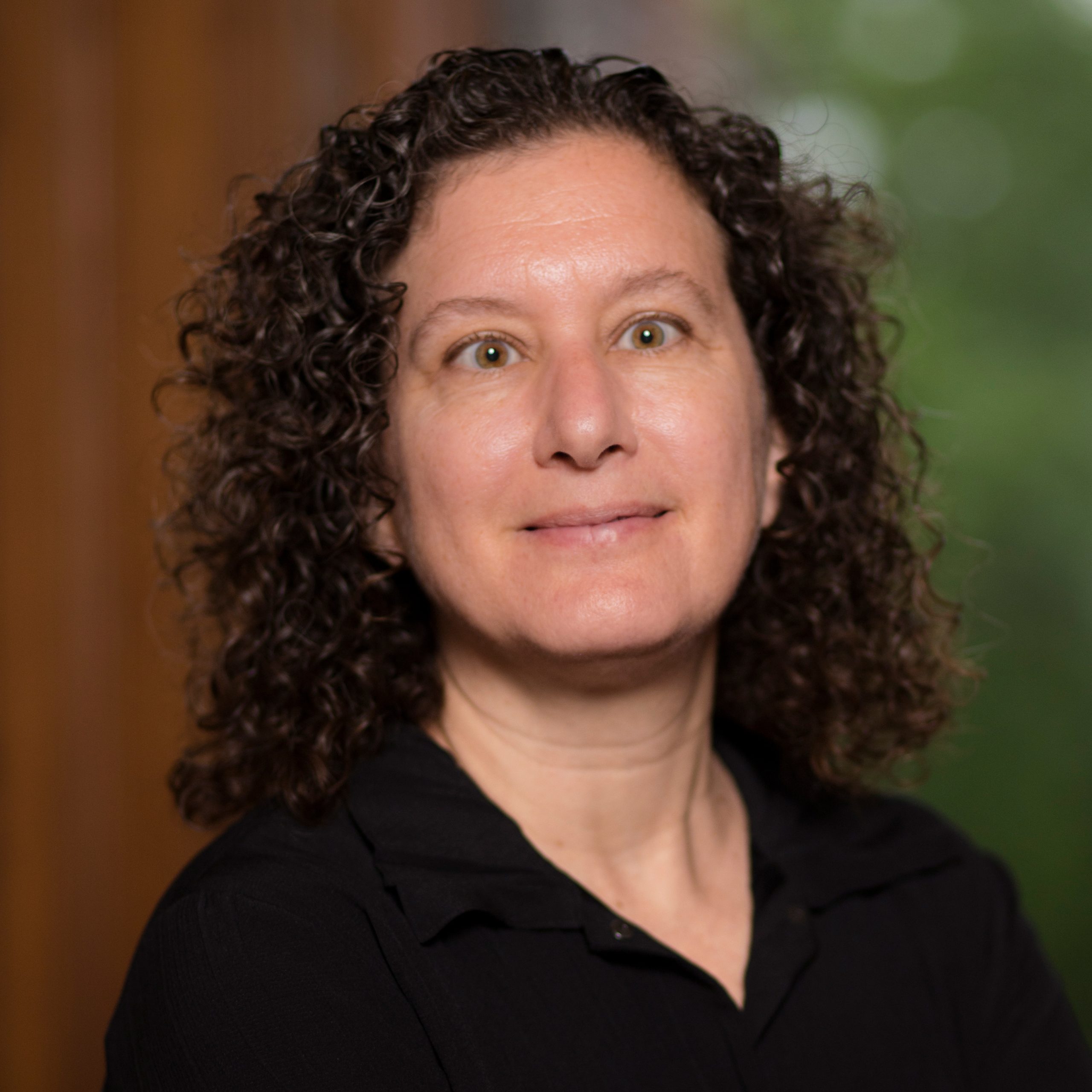 A person with curly brown hair wearing a black shirt is standing indoors. The background is softly blurred with hints of brown and green.