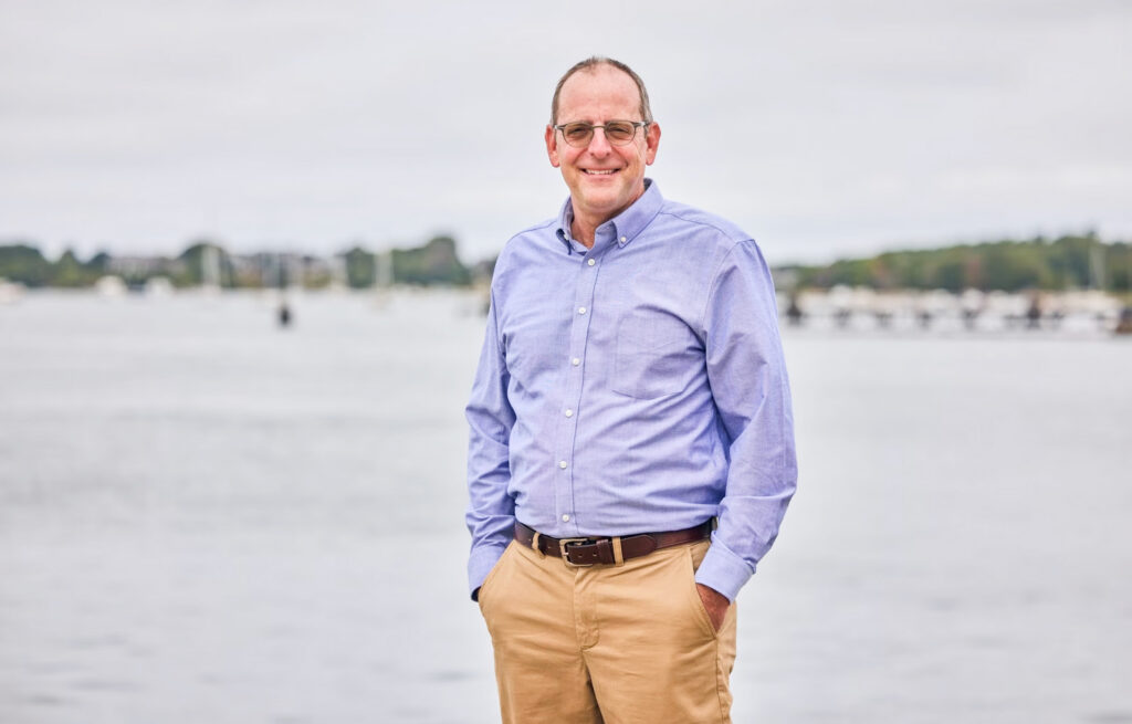 A man wearing glasses, a blue button-down shirt, and khaki pants stands by a body of water with boats and trees in the background, smiling at the camera.