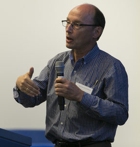 A man wearing glasses and a blue checkered shirt speaks into a microphone while gesturing with his hand during a presentation.