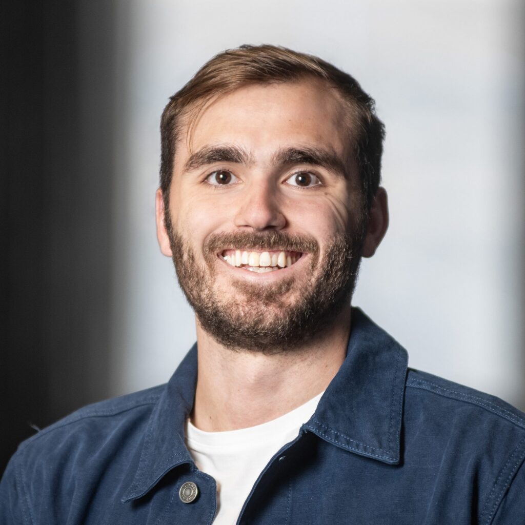 A man with short brown hair and a beard smiles at the camera. He is wearing a white shirt and a blue jacket, with a blurred background behind him.