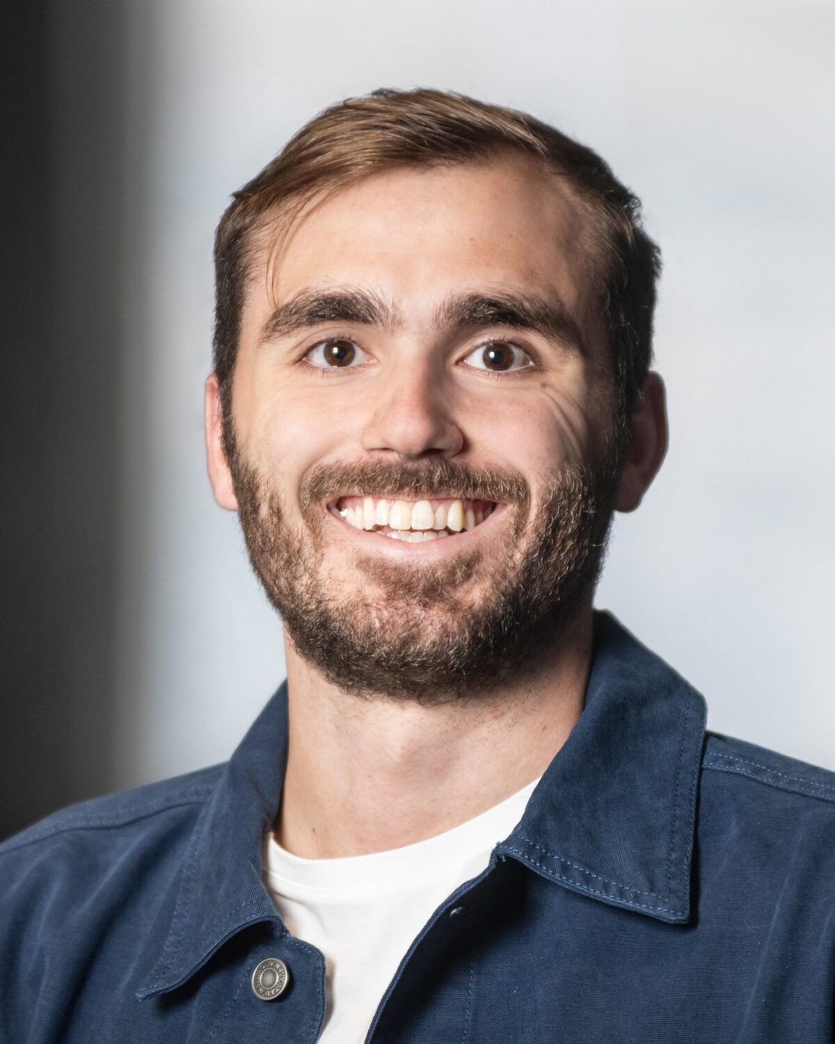 A man with short brown hair and a beard smiles at the camera. He is wearing a white shirt and a blue jacket, with a blurred background behind him.