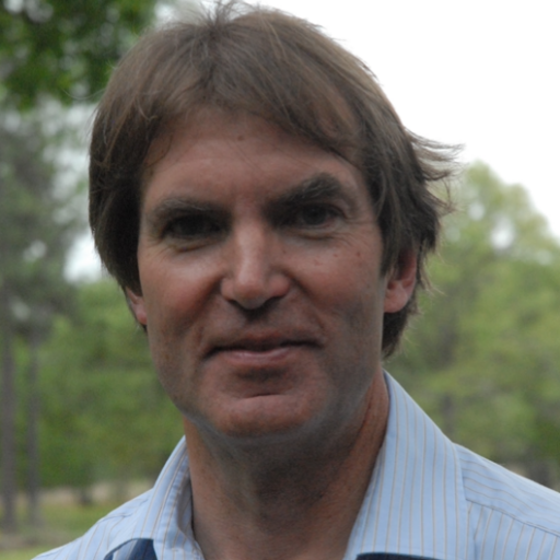 A man with brown hair, wearing a light blue striped shirt, stands outdoors with trees and greenery blurred in the background.