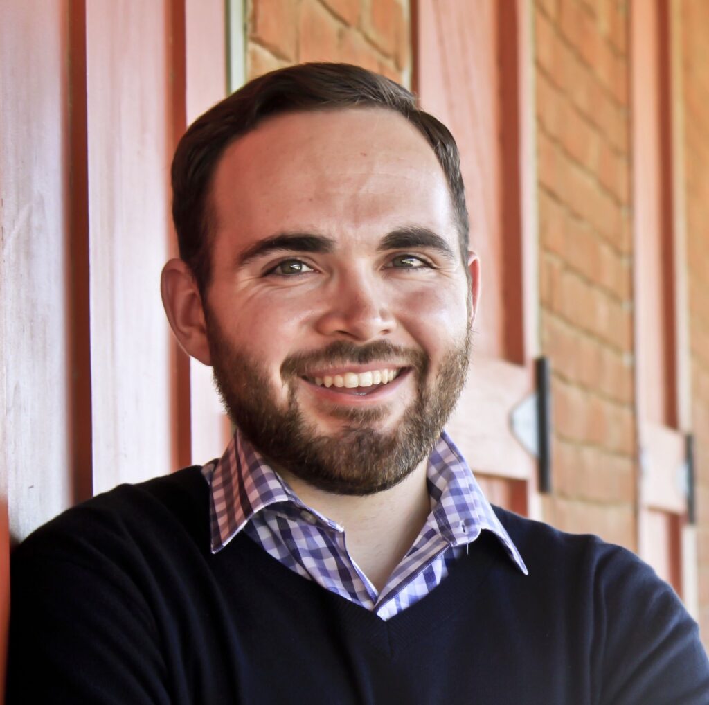A man with short brown hair and a beard, wearing a navy sweater over a checked shirt, smiles while leaning against a red wooden wall with brick accents.