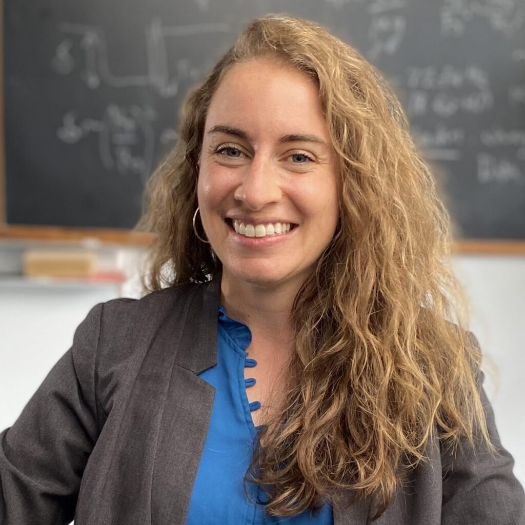 A woman with long curly hair, wearing a blue blouse and gray blazer, smiles while sitting in front of a chalkboard filled with equations.