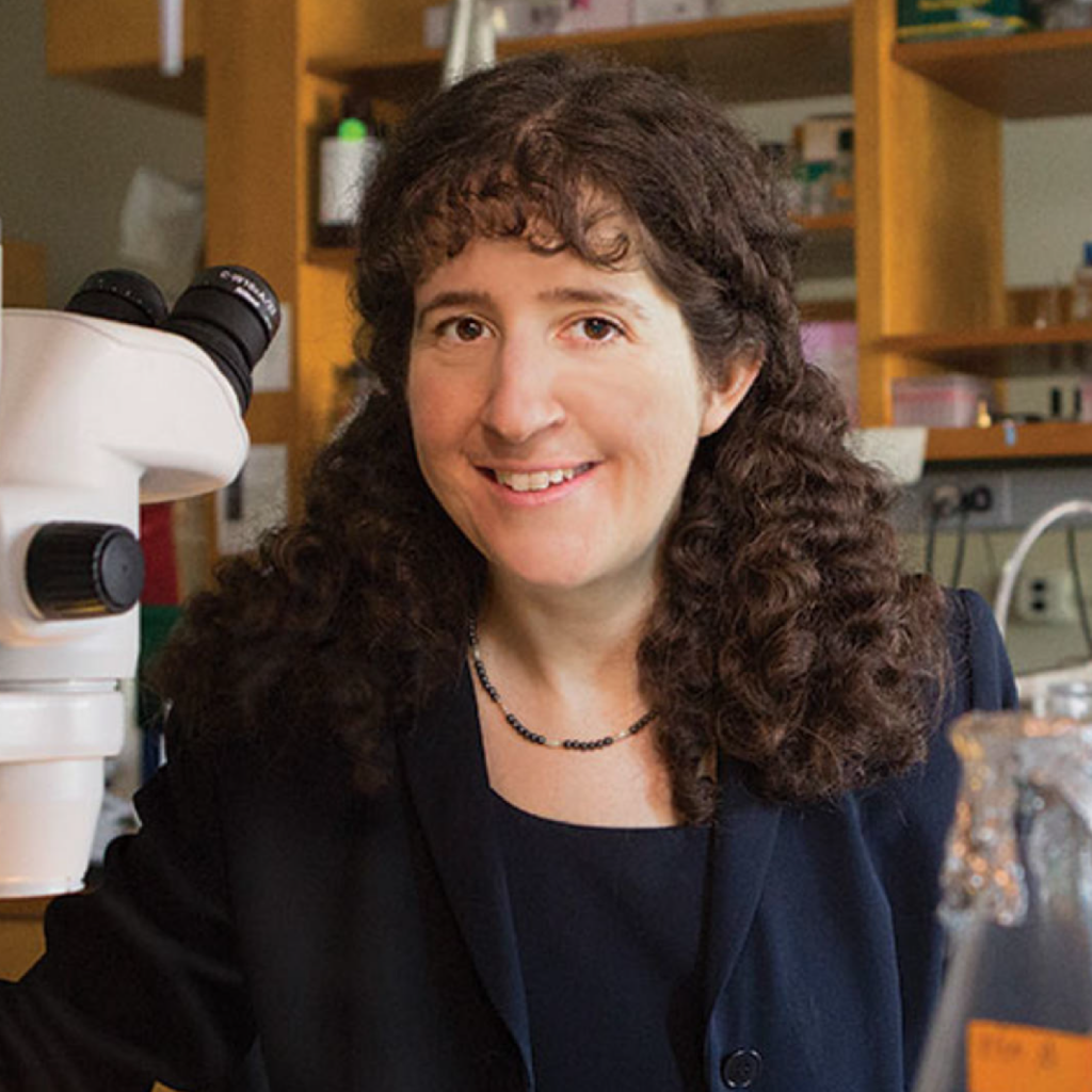 A woman with curly brown hair smiles in a laboratory, standing beside a microscope and lab equipment. She is wearing a dark blazer and a necklace. Shelves with lab supplies are visible in the background.