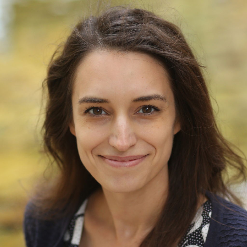 A woman with long brown hair and a slight smile is wearing a dark sweater over a patterned top, standing outdoors with a blurred yellow-green background.