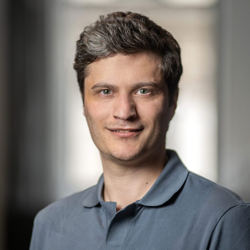 A man with short, wavy brown hair and hazel eyes, wearing a gray collared shirt, smiles softly at the camera. The background is blurred and softly lit.