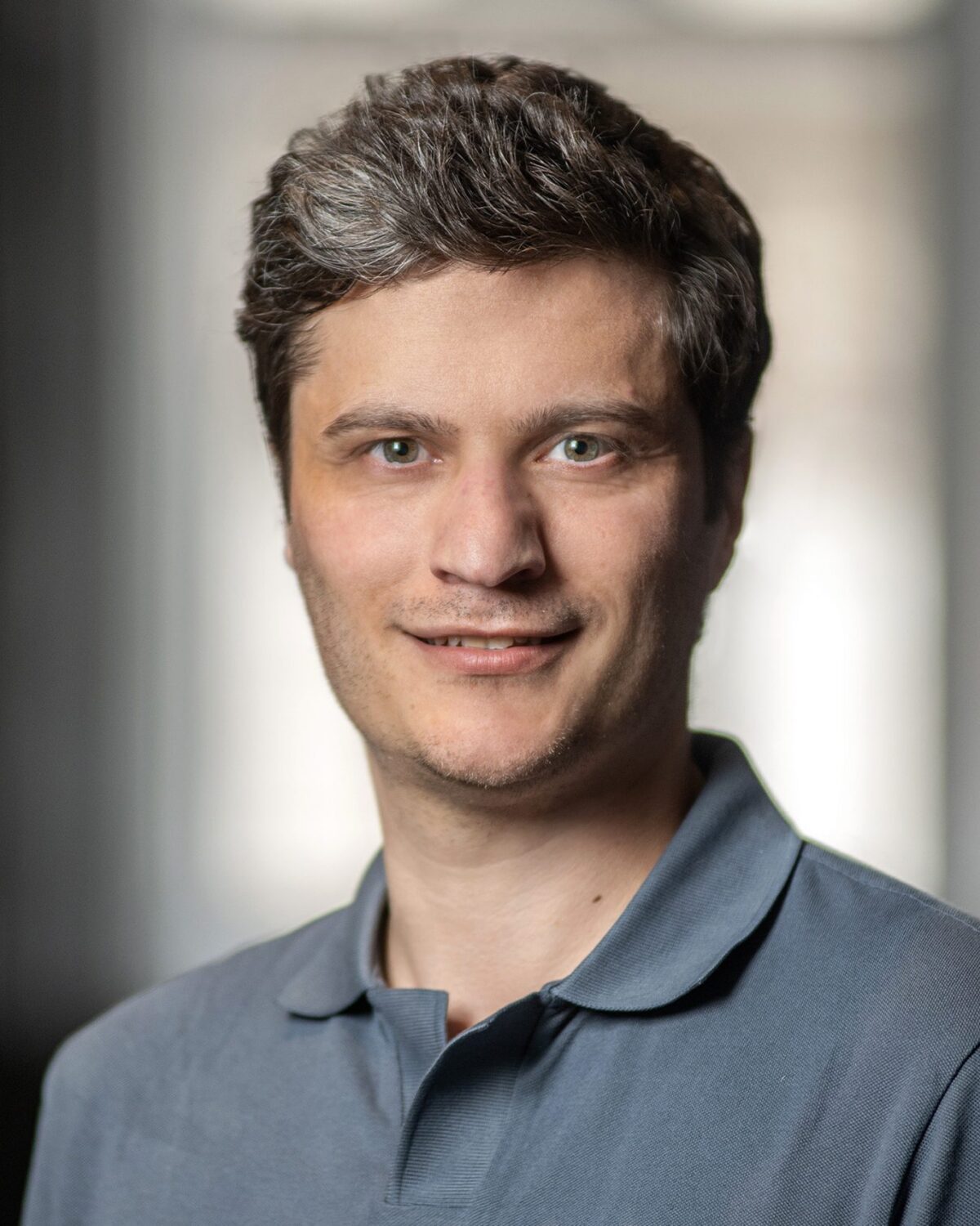A man with short, wavy brown hair and hazel eyes, wearing a gray collared shirt, smiles softly at the camera. The background is blurred and softly lit.