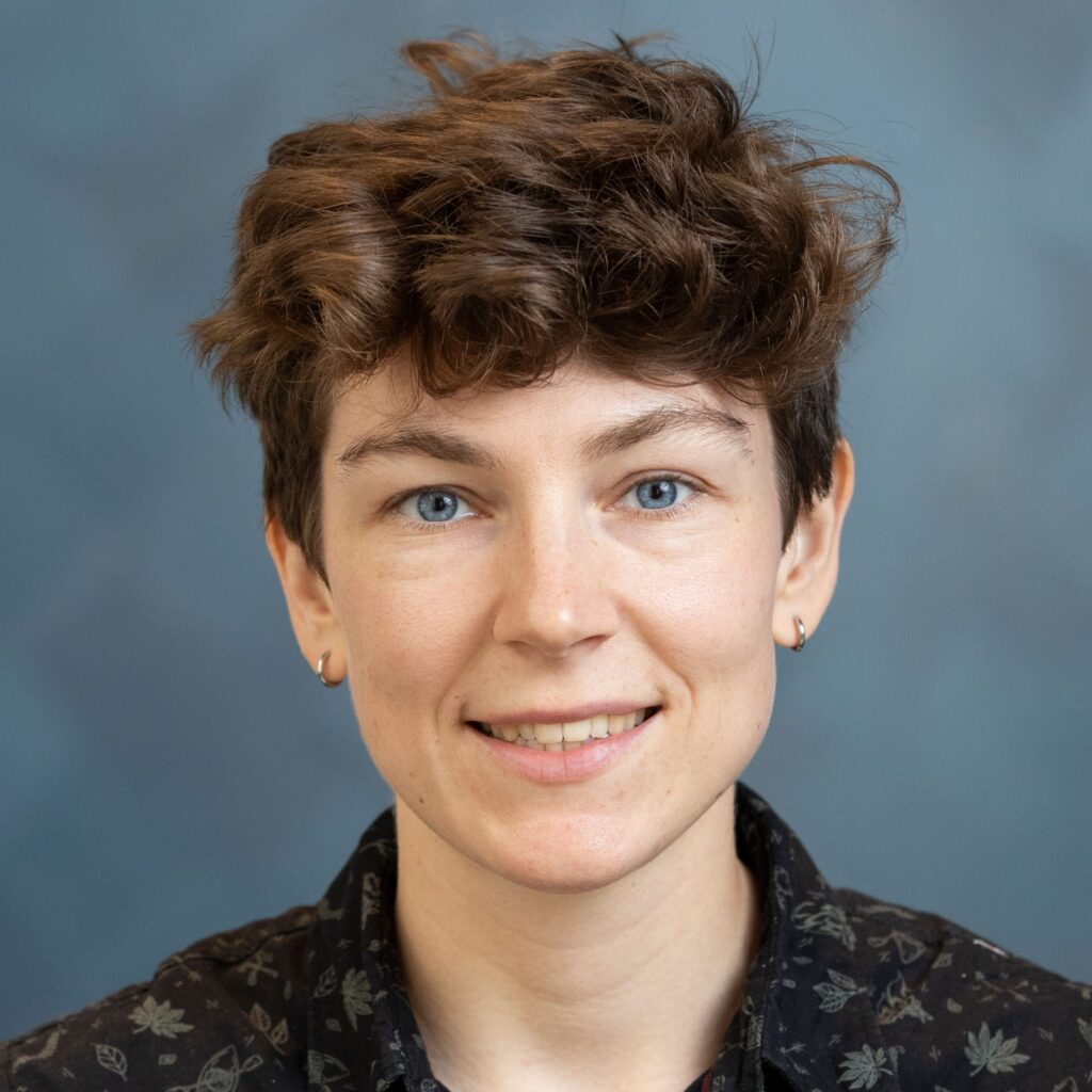 Person with short, curly brown hair and blue eyes smiling at the camera, wearing small hoop earrings and a dark shirt with a subtle pattern, against a plain blue-grey background.