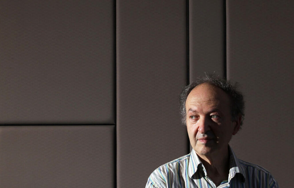 A man with thinning hair and a striped shirt stands in front of a padded wall, looking slightly to the right. Light from the left highlights his face while the background remains in shadow.